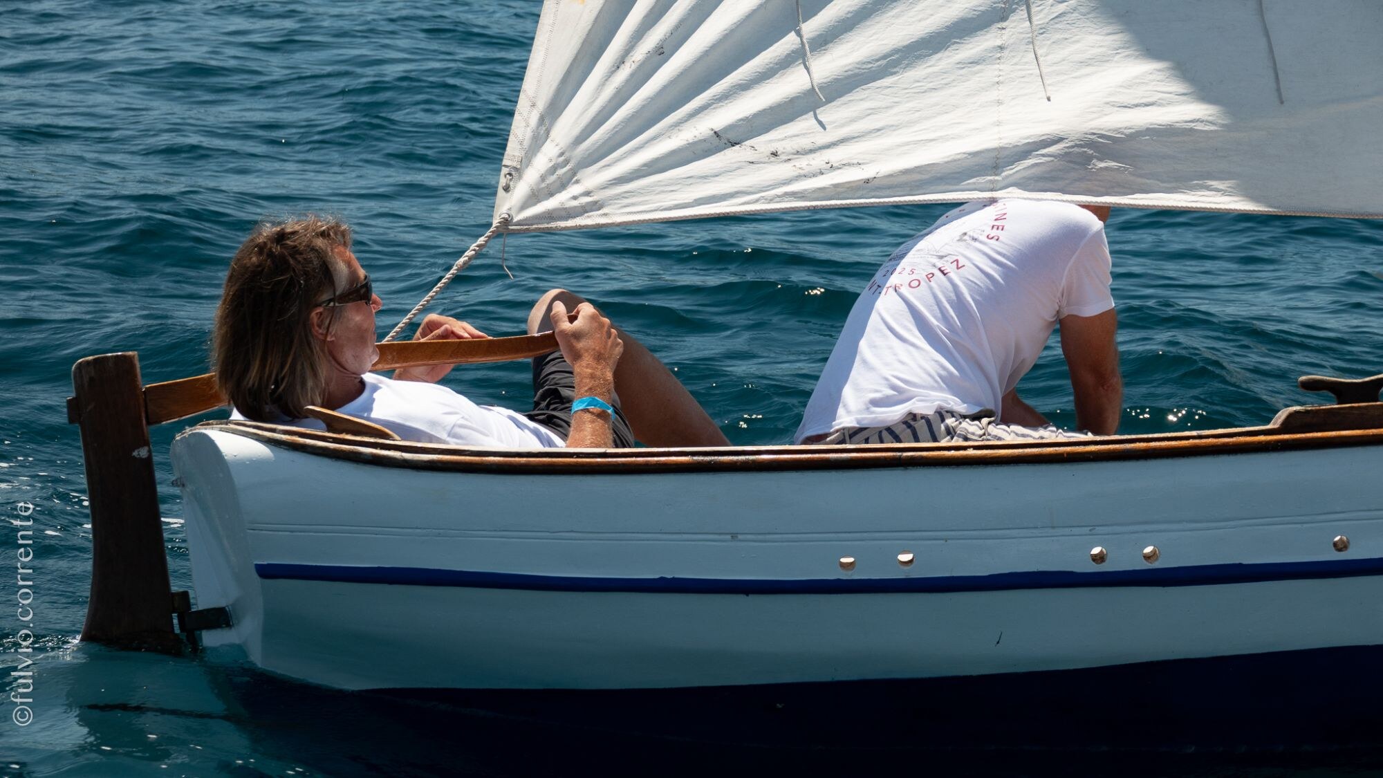 Deux équipiers à bord d’une petite barque blanche à liseré bleu, voile blanche.