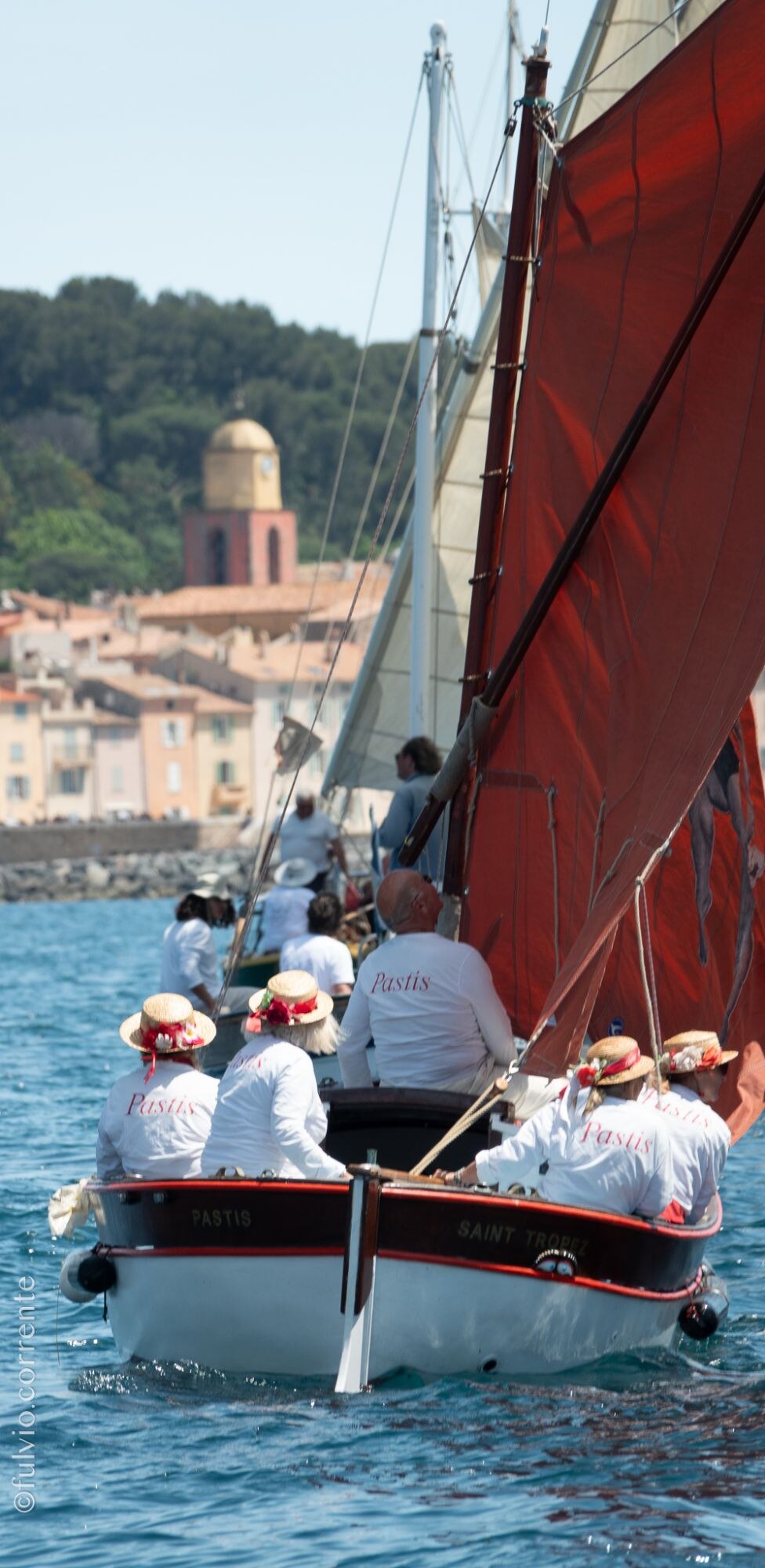 Barque blanche et rouge à voile rousse, équipage en blanc et chapeaux de paille, clocher derrière.
