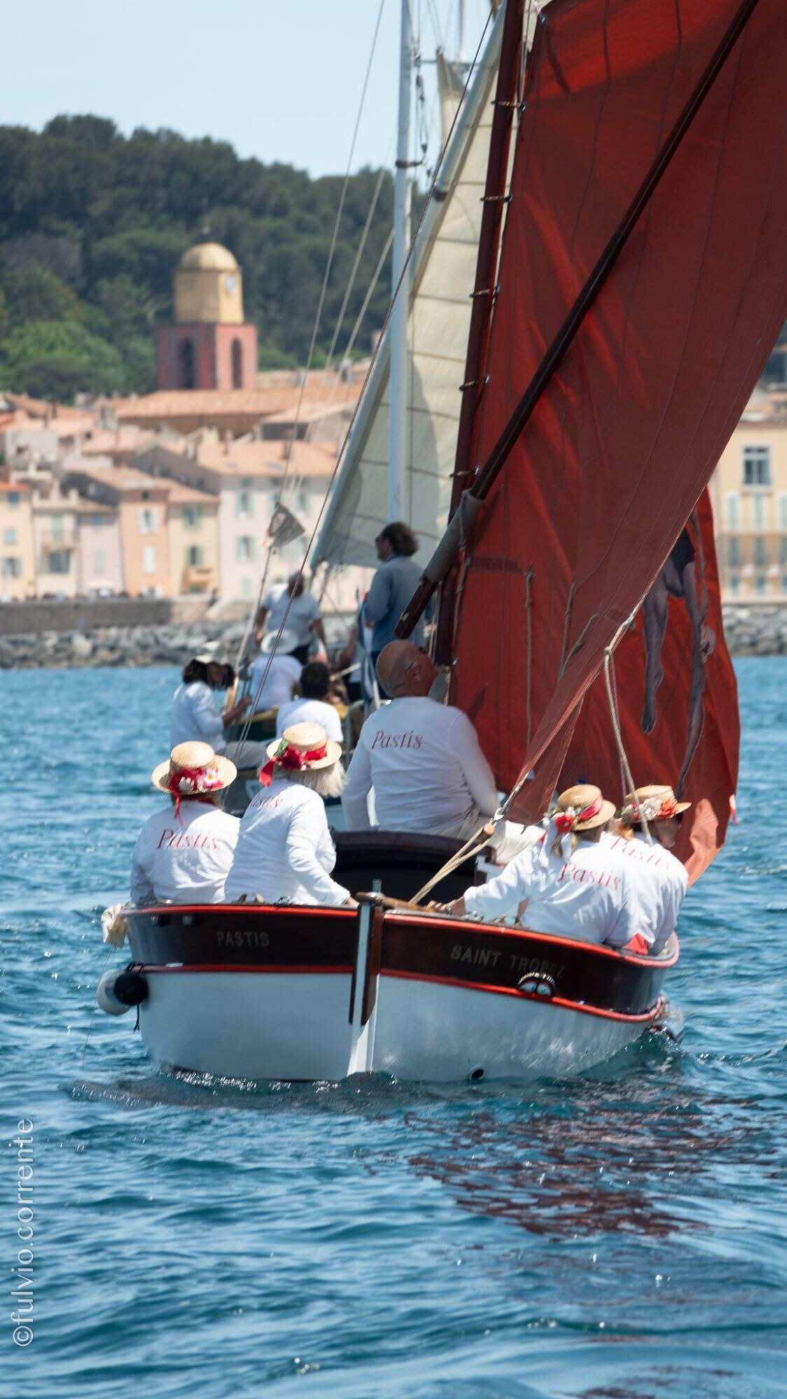 Barque à voile rousse, équipage en blanc et chapeaux de paille, village en arrière-plan.