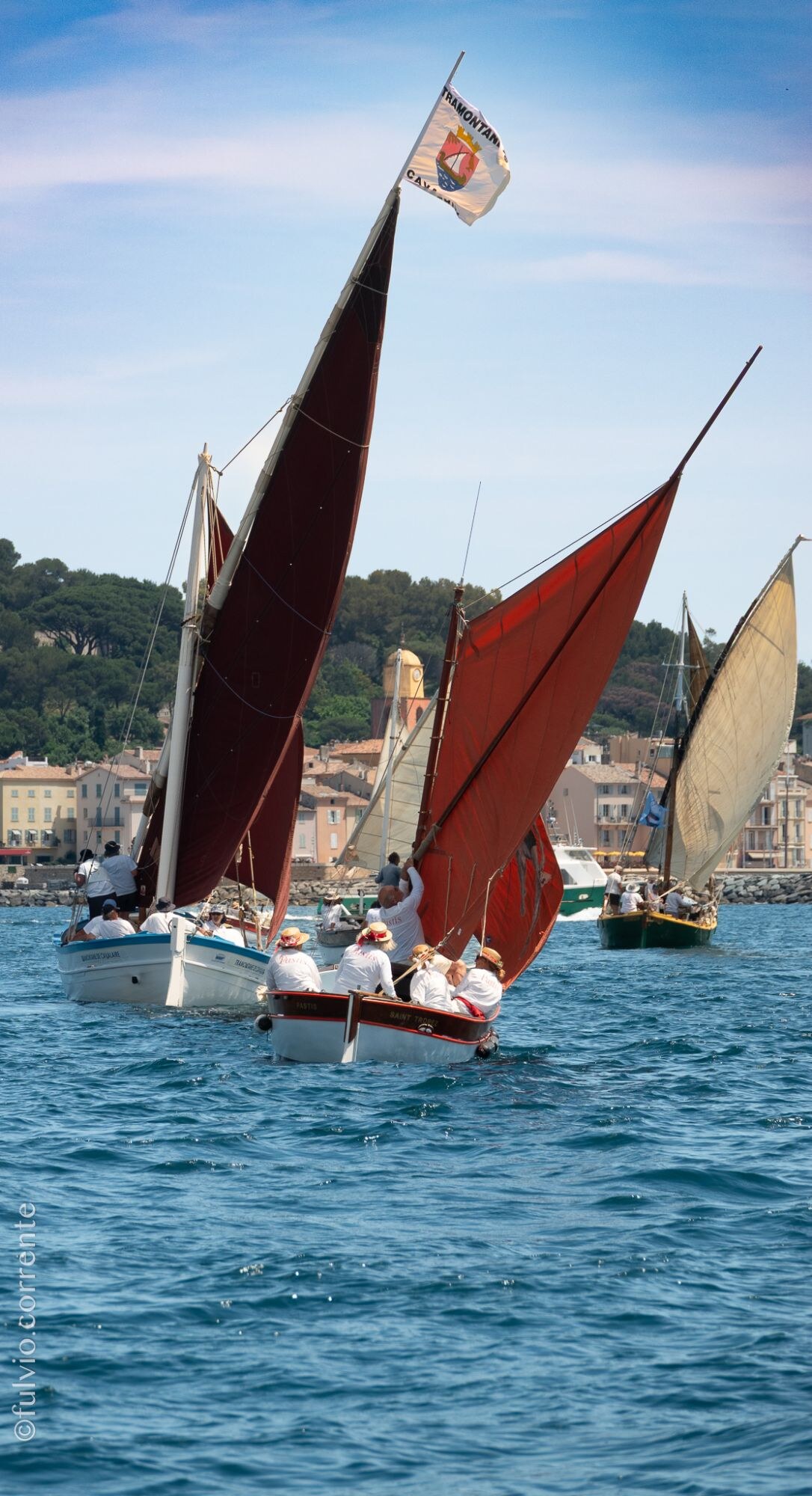 Voiles latines rousses de plusieurs barques, pavillon en haut du mât, village derrière.
