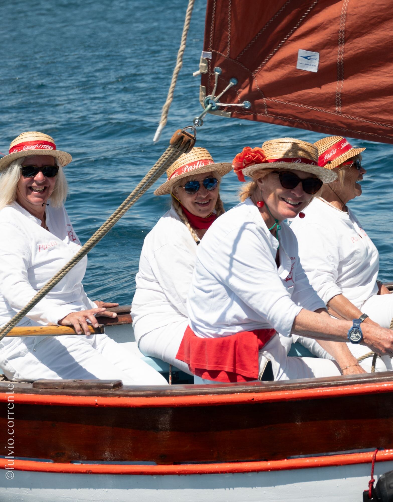 Équipières souriantes en blanc et chapeaux de paille à bord d’une barque rouge et blanche.