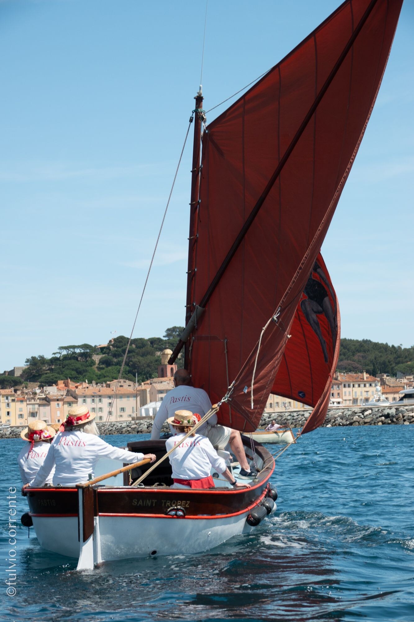 Barque à voile rousse avec équipage en chapeaux de paille, village au fond.