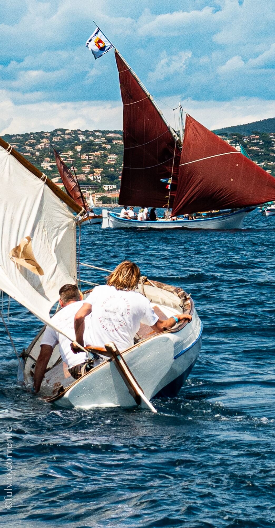 Équipiers manœuvrant une barque blanche au premier plan, voile rousse derrière.