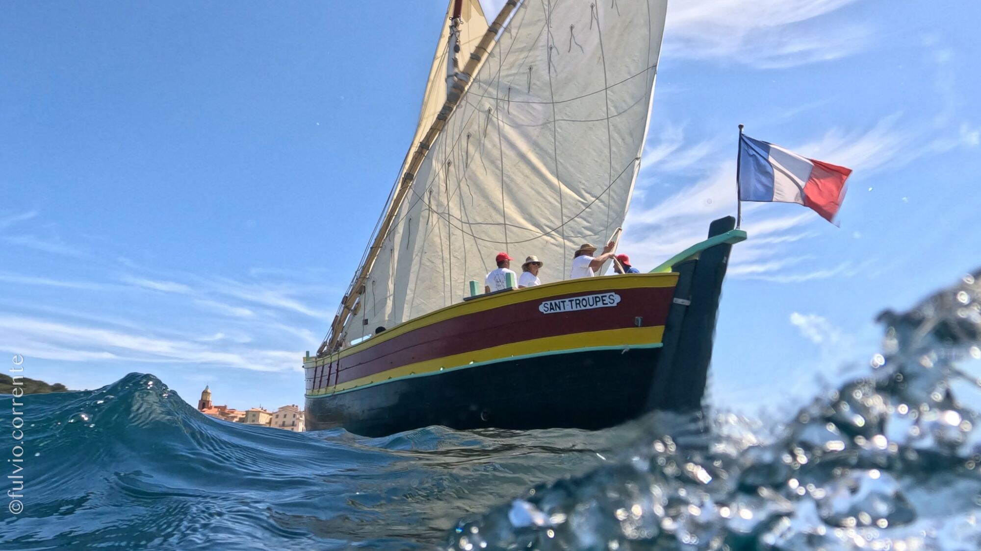 Barque à coque sombre et drapeau tricolore, voile blanche, vagues au premier plan.