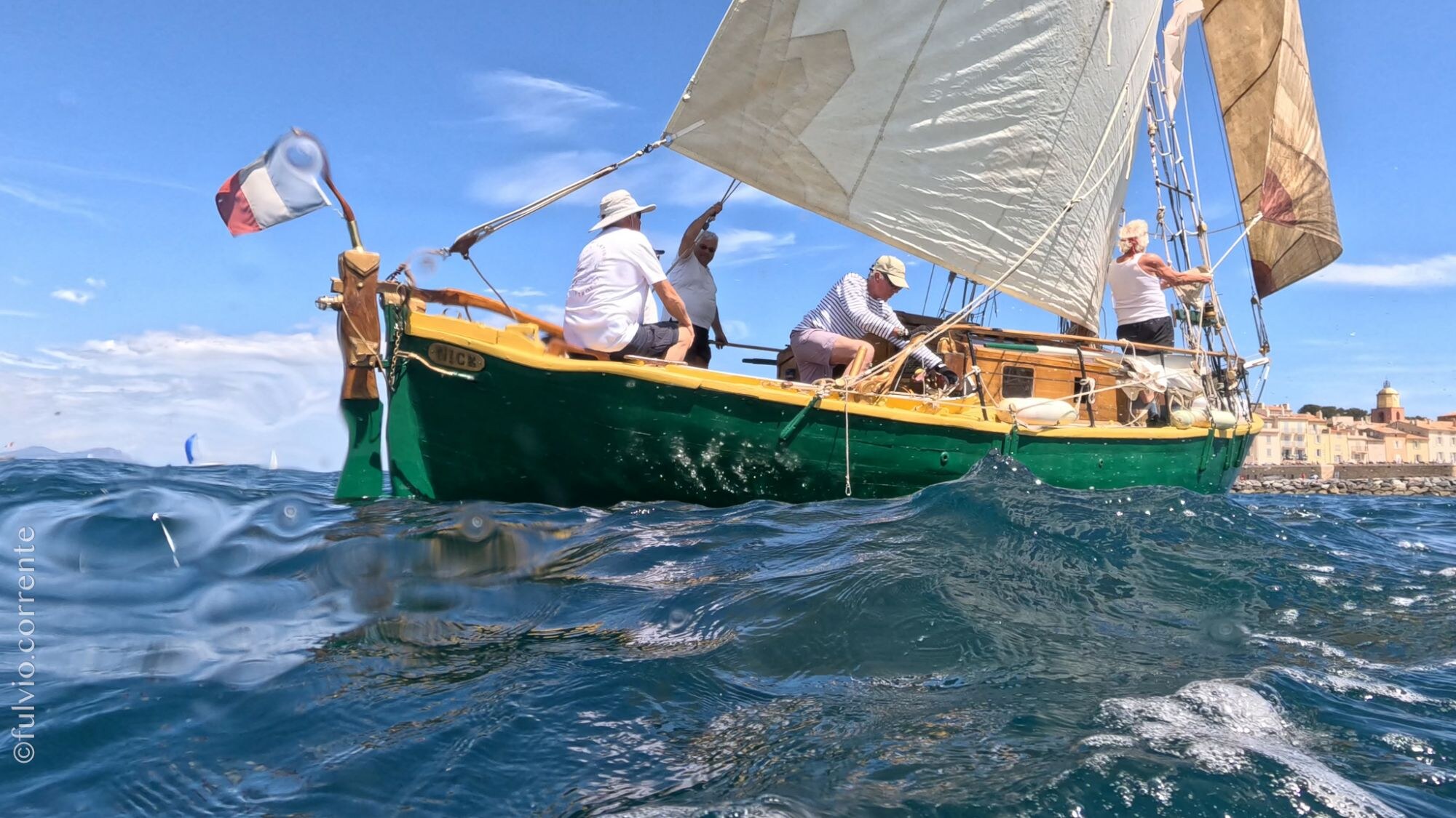 Barque à coque verte et jaune sous voile claire, équipage à bord, village au fond.