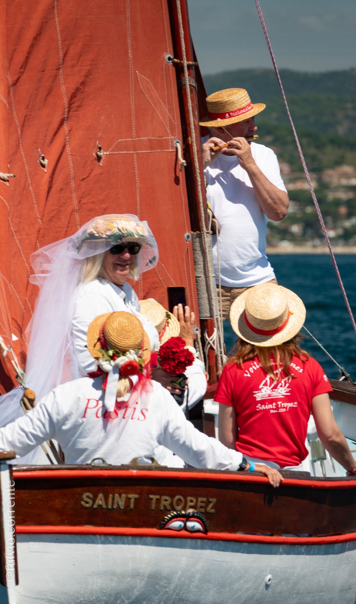 Équipière en tenue de mariée voilée et chapeau de paille, autres équipiers à bord d’une barque rouge et blanche.