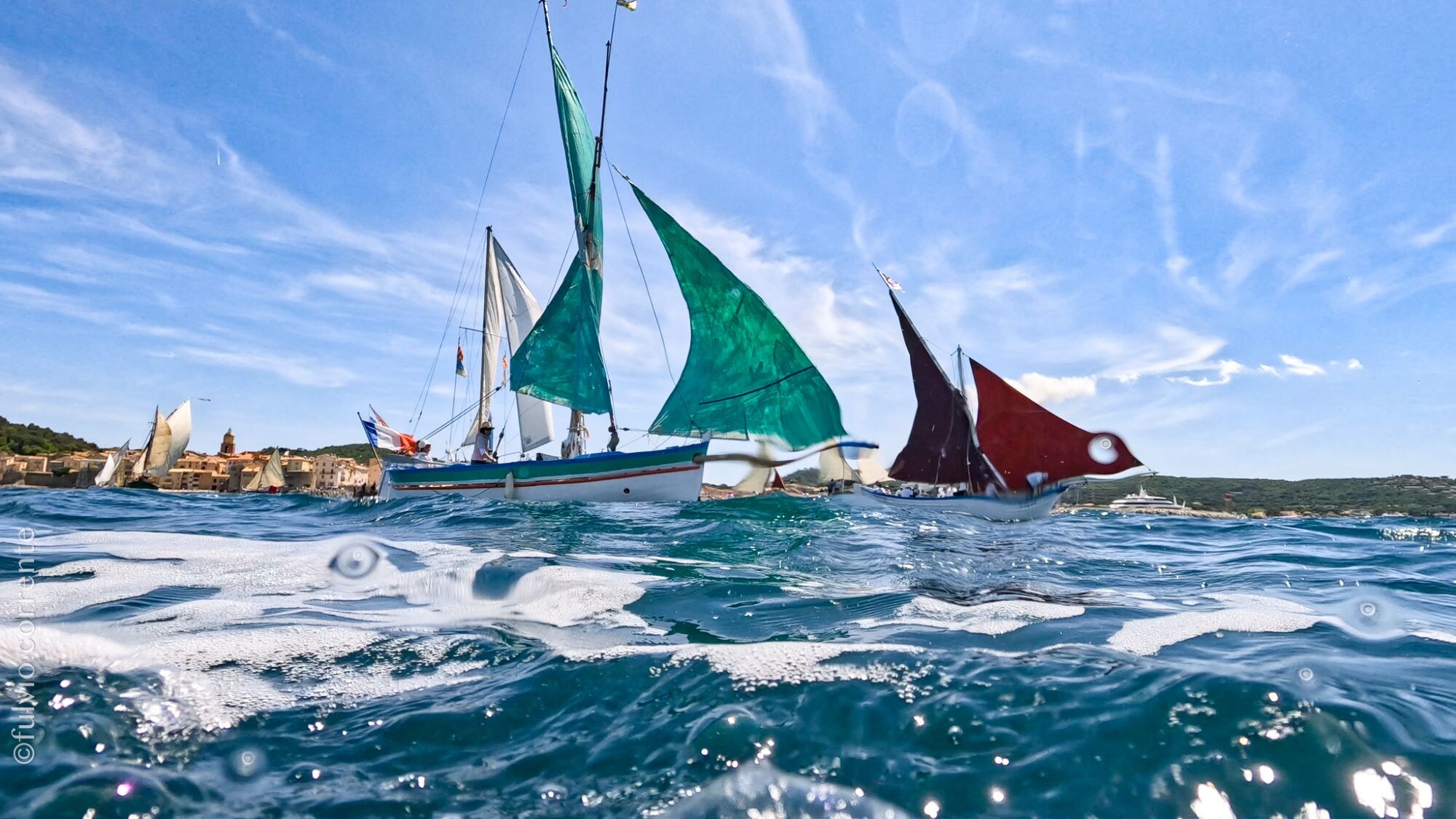 Barques à voiles vertes et brunes vues à fleur d’eau, côte au loin.