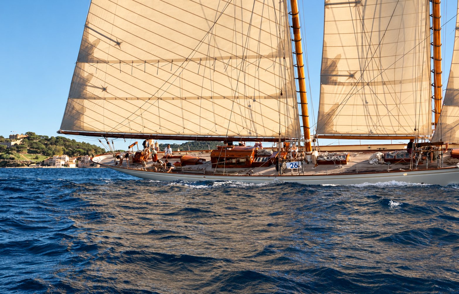 Goélette classique aux voiles beiges naviguant près d’une côte méditerranéenne.