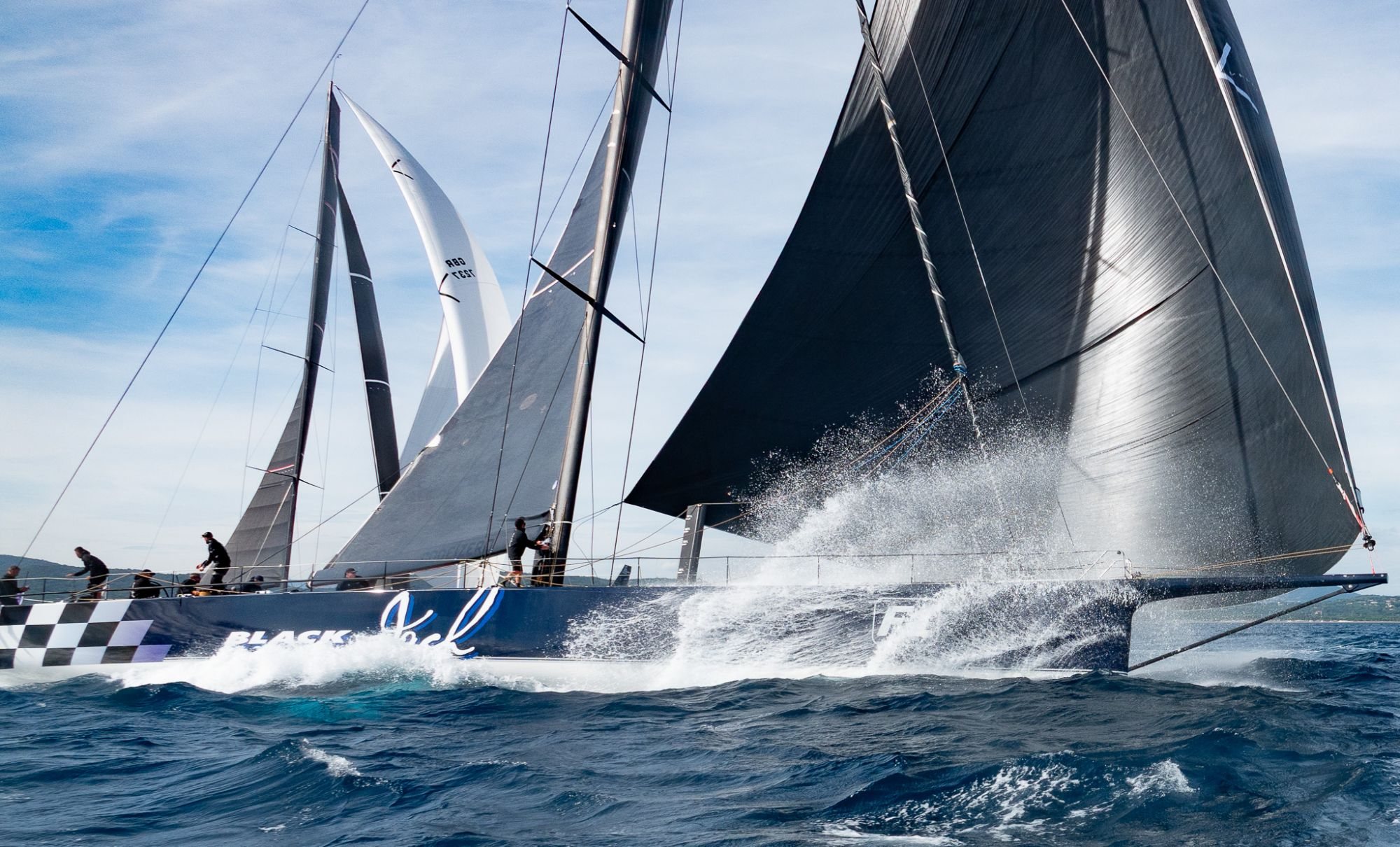 Voilier de course noir fendant les vagues sous un ciel bleu.